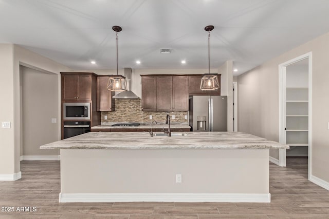 kitchen featuring wall chimney range hood, sink, appliances with stainless steel finishes, a kitchen island with sink, and dark brown cabinetry