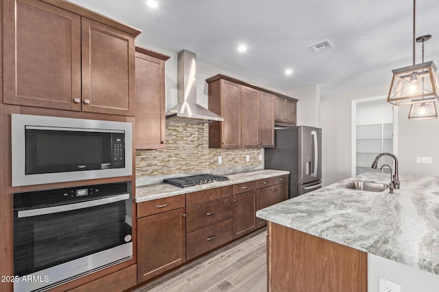 kitchen with sink, hanging light fixtures, stainless steel appliances, light stone countertops, and wall chimney range hood