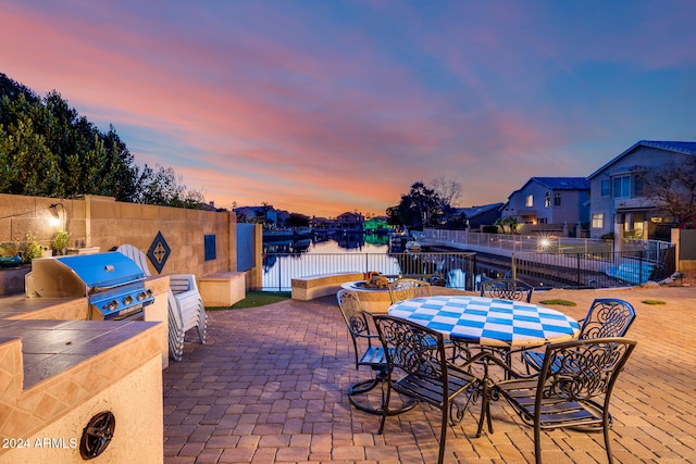 patio terrace at dusk with an outdoor kitchen, a grill, and a water view