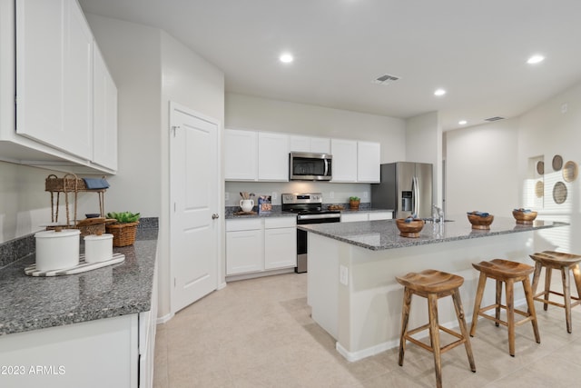 kitchen with dark stone counters, white cabinets, a center island with sink, appliances with stainless steel finishes, and a breakfast bar area