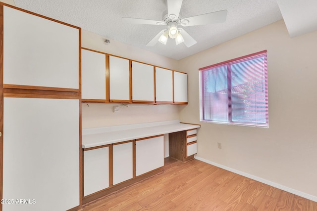 unfurnished office with a textured ceiling, a ceiling fan, baseboards, light wood-style floors, and built in study area