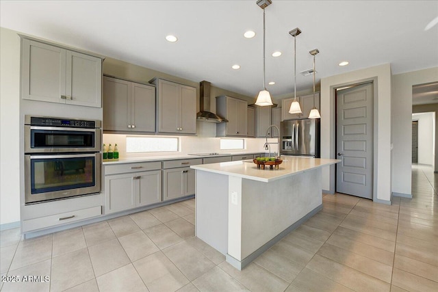 kitchen featuring pendant lighting, wall chimney range hood, gray cabinets, a kitchen island with sink, and stainless steel appliances