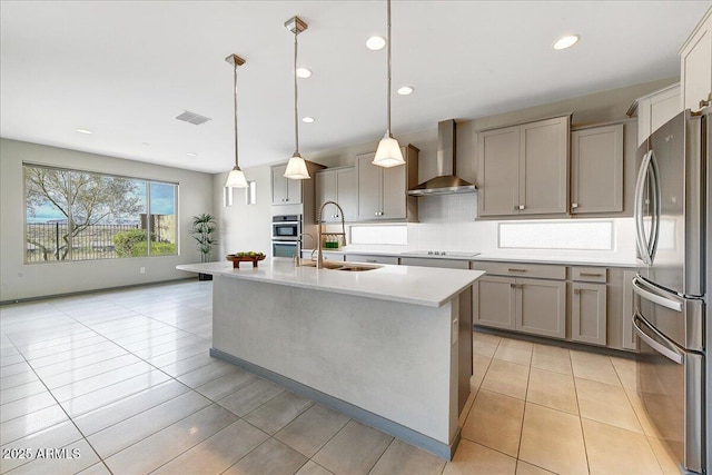 kitchen featuring wall chimney exhaust hood, sink, appliances with stainless steel finishes, pendant lighting, and a kitchen island with sink