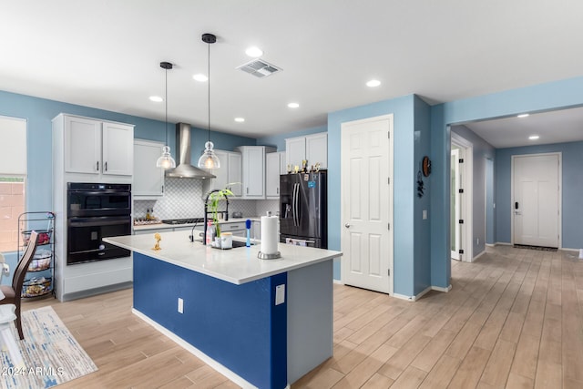 kitchen featuring black appliances, a center island with sink, backsplash, light wood-type flooring, and wall chimney exhaust hood