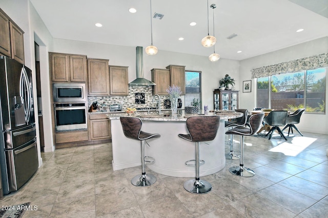 kitchen featuring light stone counters, wall chimney exhaust hood, stainless steel appliances, decorative light fixtures, and a center island with sink