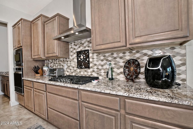 kitchen with wall chimney range hood, decorative backsplash, light tile patterned flooring, light stone counters, and stainless steel appliances