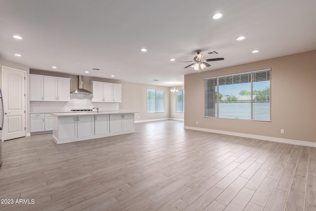 kitchen featuring a kitchen island with sink, ceiling fan with notable chandelier, light hardwood / wood-style flooring, wall chimney exhaust hood, and white cabinetry