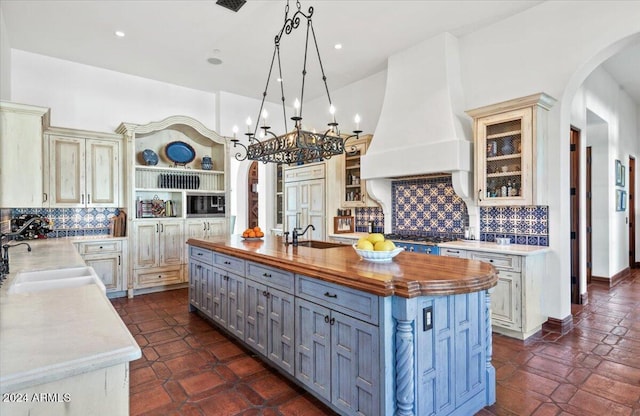 kitchen with pendant lighting, premium range hood, sink, an island with sink, and butcher block countertops