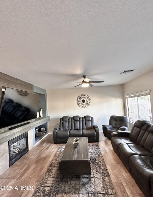 living room with wood finished floors, ceiling fan, a fireplace, visible vents, and a textured ceiling
