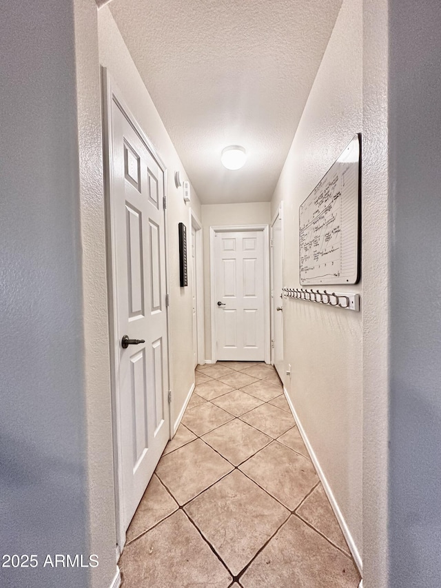 hall featuring light tile patterned floors, a textured ceiling, a textured wall, and baseboards