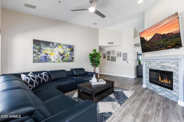 living room featuring a ceiling fan, wood finished floors, visible vents, baseboards, and a stone fireplace