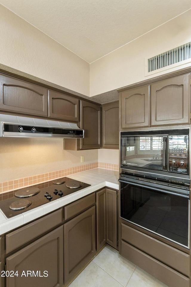 kitchen featuring tile counters, exhaust hood, dark brown cabinets, light tile patterned floors, and black appliances