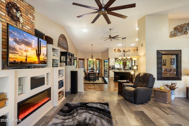 living area featuring visible vents, wood finished floors, high vaulted ceiling, ceiling fan with notable chandelier, and a glass covered fireplace