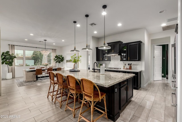 1931 E Aster Pl, Chandler, AZ 85286 - kitchen with dark cabinetry, a large island with sink, decorative backsplash, a kitchen breakfast bar, and light stone countertops