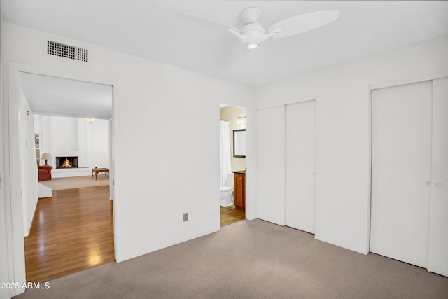 unfurnished bedroom with multiple closets, a warm lit fireplace, carpet flooring, connected bathroom, and ceiling fan