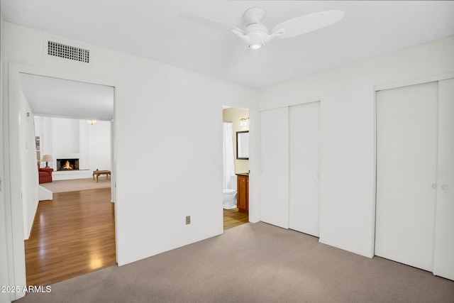 unfurnished bedroom with multiple closets, a warm lit fireplace, carpet flooring, ensuite bathroom, and ceiling fan