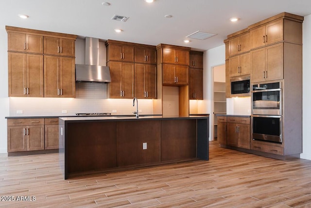 kitchen featuring wall chimney range hood, sink, built in microwave, double oven, and an island with sink