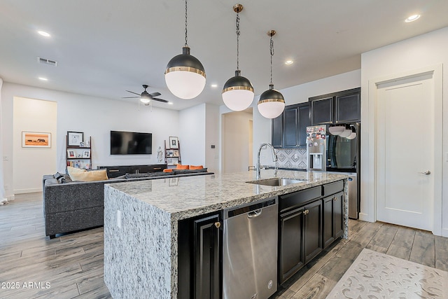 kitchen with a sink, a center island with sink, light wood-style flooring, stainless steel appliances, and dark cabinets