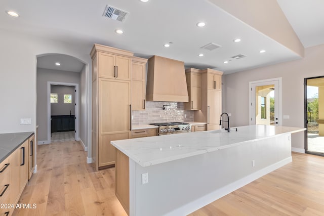 kitchen featuring high end stainless steel range oven, custom range hood, a healthy amount of sunlight, a large island with sink, and light brown cabinets