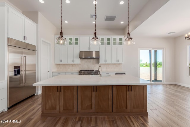 kitchen with a center island with sink, glass insert cabinets, built in refrigerator, light countertops, and under cabinet range hood