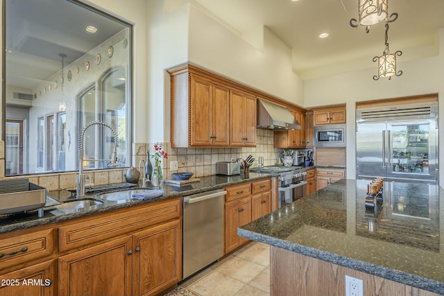 kitchen with wall chimney range hood, sink, dark stone countertops, built in appliances, and tasteful backsplash