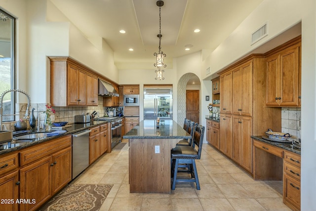 kitchen with a kitchen island, a kitchen bar, dark stone counters, built in appliances, and wall chimney exhaust hood