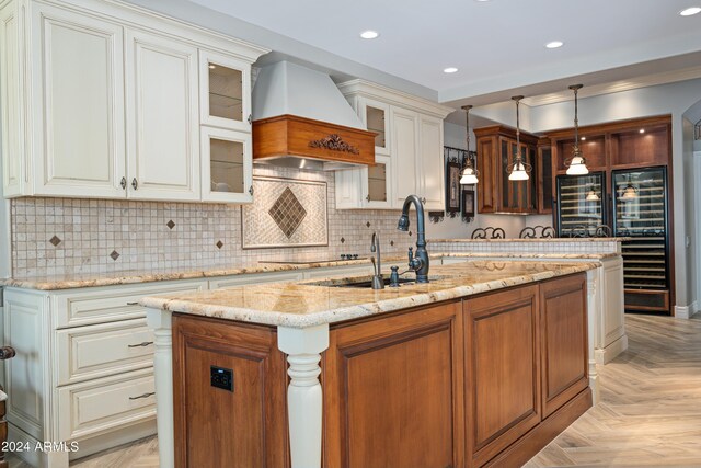 kitchen with a kitchen island with sink, backsplash, light stone counters, hanging light fixtures, and custom range hood