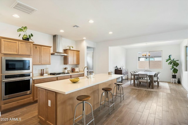 kitchen with a kitchen bar, appliances with stainless steel finishes, a kitchen island with sink, wall chimney range hood, and an inviting chandelier