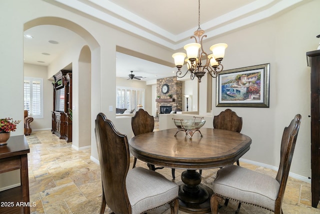 dining area featuring ceiling fan with notable chandelier, a raised ceiling, and a stone fireplace