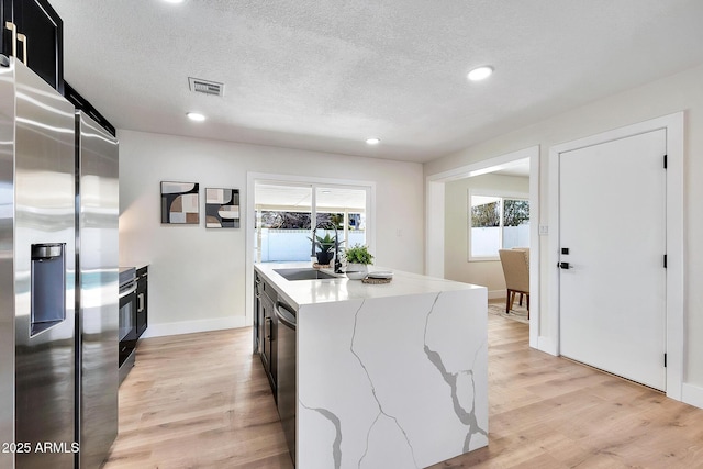 kitchen featuring a textured ceiling, stainless steel appliances, a sink, light wood-type flooring, and a center island with sink