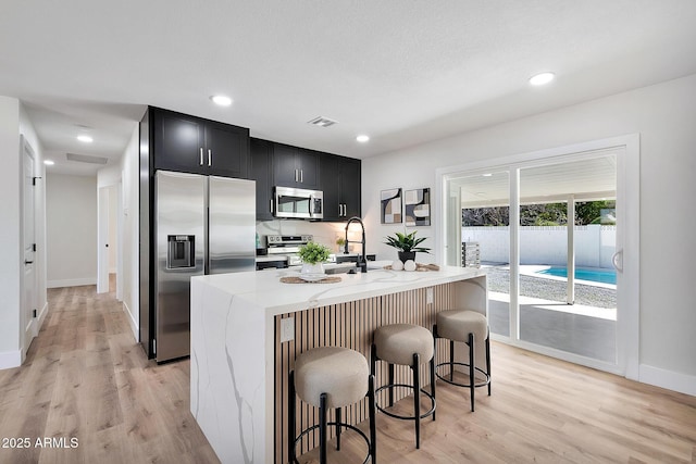 kitchen with a center island with sink, appliances with stainless steel finishes, light wood-style floors, light stone countertops, and dark cabinets