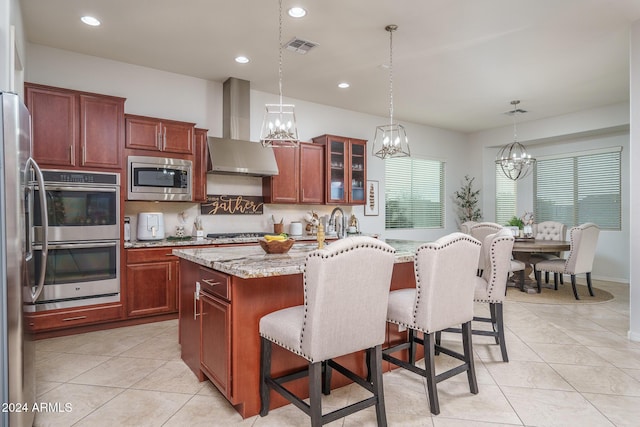 kitchen with pendant lighting, a kitchen island with sink, wall chimney exhaust hood, appliances with stainless steel finishes, and light stone counters