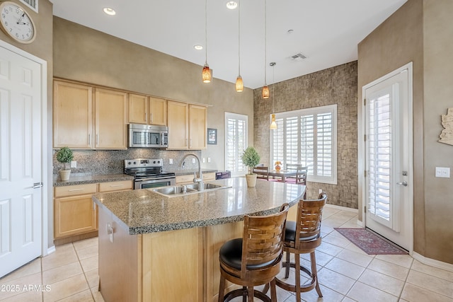 kitchen featuring light stone counters, decorative light fixtures, a center island with sink, light brown cabinets, and appliances with stainless steel finishes