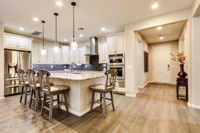 kitchen with light stone countertops, wall chimney exhaust hood, stainless steel appliances, an island with sink, and pendant lighting