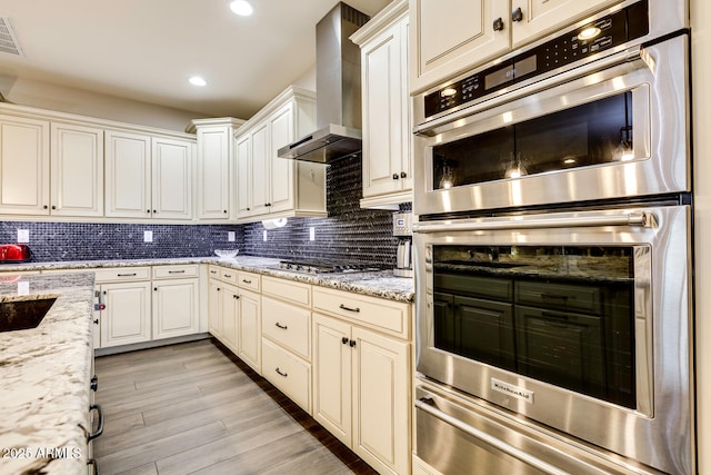 kitchen featuring light stone counters, wall chimney range hood, stainless steel appliances, and tasteful backsplash