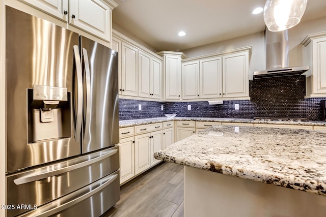 kitchen with light stone countertops, wall chimney range hood, gas stovetop, stainless steel fridge, and pendant lighting