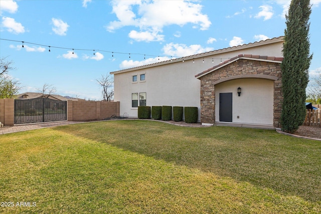 rear view of house featuring fence, stucco siding, a yard, stone siding, and a gate