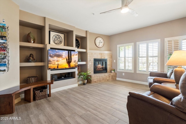 living room featuring built in shelves, wood finished floors, baseboards, a ceiling fan, and a tiled fireplace