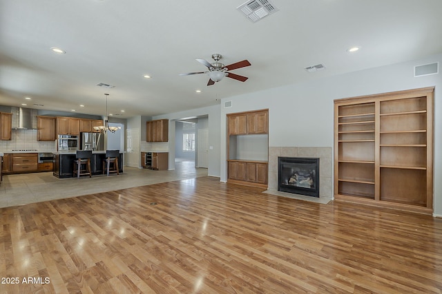 unfurnished living room with a tiled fireplace, light wood-style flooring, ceiling fan with notable chandelier, and visible vents