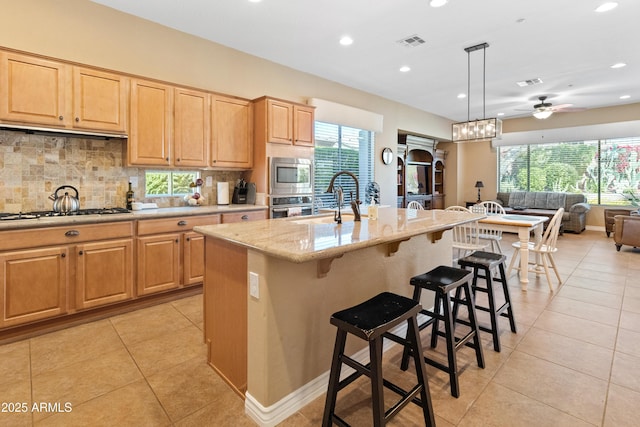 kitchen with tasteful backsplash, hanging light fixtures, an island with sink, stainless steel appliances, and light stone countertops