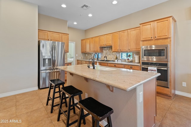 kitchen featuring backsplash, stainless steel appliances, light stone countertops, an island with sink, and light brown cabinetry