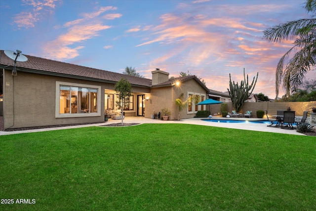 back of property at dusk with a lawn, a chimney, fence, a patio area, and stucco siding