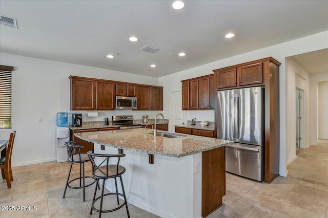 kitchen with a kitchen bar, sink, an island with sink, stainless steel appliances, and light stone countertops