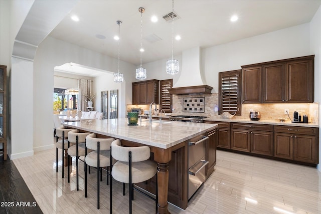 kitchen featuring light stone countertops, hanging light fixtures, tasteful backsplash, premium range hood, and a center island with sink