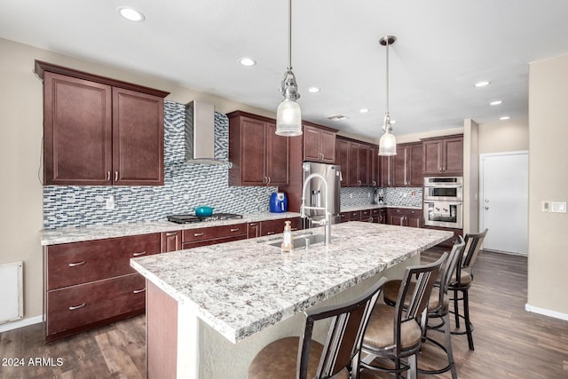 kitchen featuring a center island with sink, wall chimney exhaust hood, pendant lighting, and appliances with stainless steel finishes