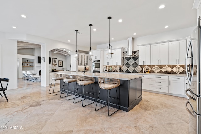 kitchen featuring a large island with sink, wall chimney range hood, decorative light fixtures, and white cabinetry