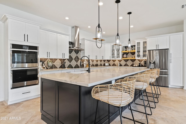 kitchen with a large island with sink, wall chimney range hood, hanging light fixtures, white cabinetry, and appliances with stainless steel finishes