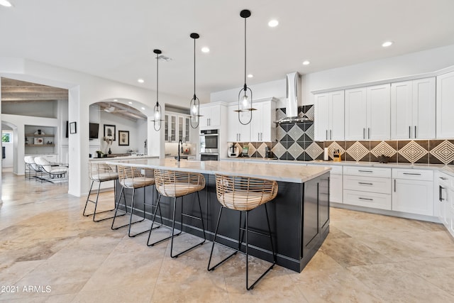 kitchen with wall chimney range hood, hanging light fixtures, white cabinetry, and a large island