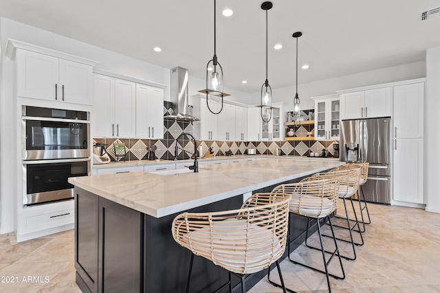 kitchen featuring a large island with sink, light stone counters, white cabinets, hanging light fixtures, and appliances with stainless steel finishes