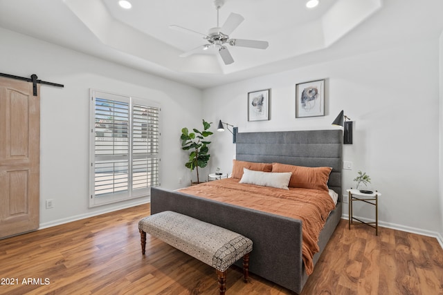 bedroom with ceiling fan, a barn door, a tray ceiling, and wood-type flooring